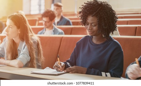 Beautiful Black Female Student Sitting Among Her Fellow Students in the Classroom, She's Writing in the Notebook and Listens to a Lecture.