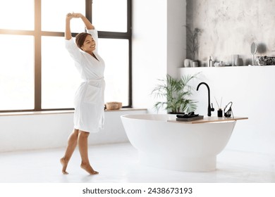 Beautiful Black Female In White Bathrobe Relaxing And Stretching In Modern Bathroom, Happy African American Woman Raising Hands Up And Smiling, Enjoying Morning Time At Home, Copy Space - Powered by Shutterstock