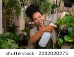 Beautiful black florist taking care of plants while spraying it with water. African american owner working and spraying water plants in store. Happy and smiling florist watering plants in shop.