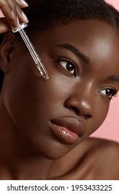 Beautiful black girl dropping serum collagen moisturizer on face. Serious young woman. Concept of face skin care. Isolated on pink background. Studio shoot