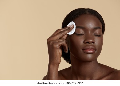 Beautiful Black Girl Touch Her Clean Face With Cotton Pad. Young Woman With Closed Eyes. Concept Of Face Skin Care. Isolated On Beige Background. Studio Shoot