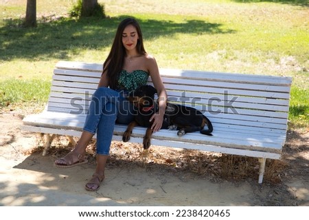 Beautiful black haired woman is sitting with her doberman puppy on a white bench in the park. The woman enjoys the company of her dog and relaxing together. Pets and animals concept.