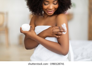 Beautiful black lady wearing towel after bath, applying cream, rubbing in nourishing body butter at home. Lovely African American woman moisturizing her skin, enjoying domestic spa procedure