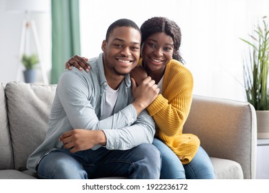 Beautiful black lovers spending time together at home while COVID-19 pandemic, happy african american couple sitting on couch in living room, embracing and looking at camera, copy space