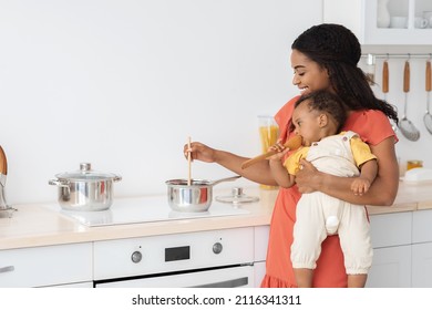 Beautiful Black Mom Cooking Food In Kitchen With Little Son In Arms, Happy African American Female Making Housework And Taking Care About Infant Baby, Preparing Healthy Meal For Family, Copy Space