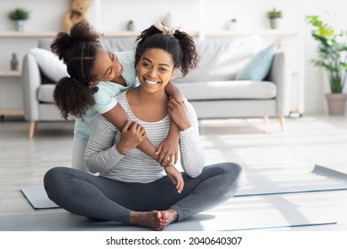 Beautiful black mother and daughter in sportswear cuddling while exercising together at home, cute teen girl hugging her pretty mom sitting on yoga mat, living room interior, copy space