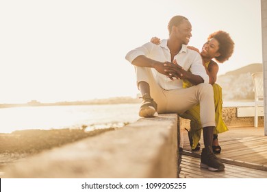 beautiful black race african couple in love and vacation sit down enjoying together with big smiles and laugh. casual clothes like fashion style with nice sunset backlight on the background. tenerife.