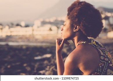 Beautiful Black Race African Model Young Woman Stay Sit Down And Think About On The Coast Near The Ocean. Sunlight On Her Face And Old Style Hair. Traditional Dress And Beauty Concept