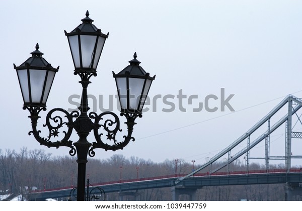 Beautiful black street lantern for three lamps, close up\
photo. Pedestrian bridge in the background. Winter morning view.\
Kyiv, Ukraine. 