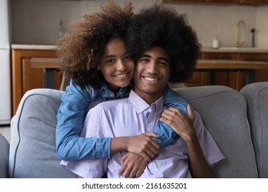 Beautiful Black Teen Girlfriend Embracing Boyfriend From Behind. Happy Cheerful African Teenage Couple In Love Enjoying Dating, Leisure At Home, Hugging, Looking At Camera, Smiling