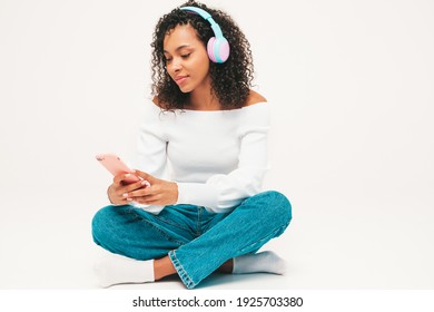 Beautiful black woman with afro curls hairstyle.Smiling model in sweater and jeans.Sexy carefree female listening music in wireless headphones.Sitting in studio on white background.Holding smartphone
