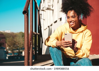 Beautiful black woman with afro curls hairstyle.Smiling model in yellow hoodie.Sexy carefree female enjoying listening music in wireless headphones.Posing on street background at sunset.Holds phone