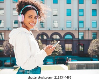 Beautiful Black Woman With Afro Curls Hairstyle.Smiling Model In White Hoodie.Sexy Carefree Female Enjoying Listening Music In Wireless Headphones.Posing On Street Background At Sunset.Holds Phone