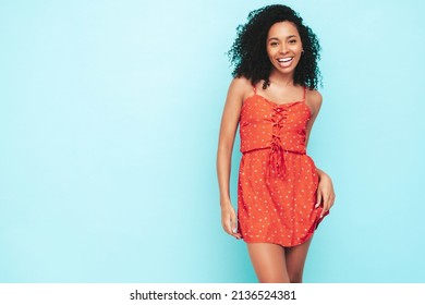 Beautiful black woman with afro curls hairstyle. Smiling model dressed in red summer dress. Sexy carefree female posing near blue wall in studio. Tanned and cheerful. At sunny day. Isolated