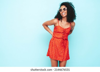 Beautiful Black Woman With Afro Curls Hairstyle. Smiling Model Dressed In Red Summer Dress. Sexy Carefree Female Posing Near Blue Wall In Studio. Tanned And Cheerful. At Sunny Day In Sunglasses