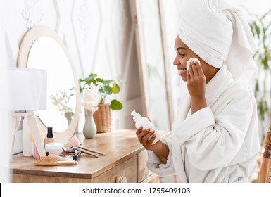 Beautiful black woman cleaning face from makeup with cleansing milk and cotton pad at home, wrapped in bathrobe and towel, sitting at dressing table - Powered by Shutterstock