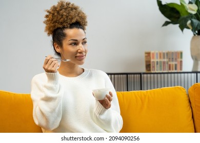 Beautiful Black Woman Eating Yogurt As Healthy Breakfast Or Snack At Home Sitting On The Couch