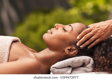 Beautiful black woman getting face massage in luxury spa. African american girl relaxing in resort spa while getting head massage. Masseuse hands massaging black woman with closed eyes at resort spa.