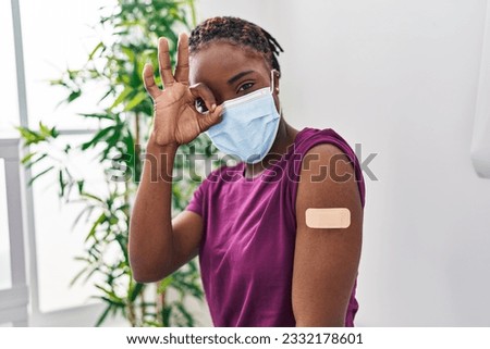 Beautiful black woman getting vaccine showing arm with band aid smiling happy doing ok sign with hand on eye looking through fingers 