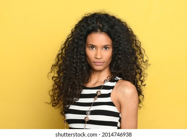 Beautiful black woman with gorgeous curly long hair posing over a yellow background looking at camera with a serious expression