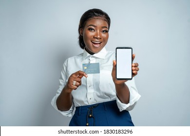 Beautiful Black Woman Happily Showing A Blank Phone Screen To Camera And Holding An ATM Or Credit Card-concept On Business And E-commerce