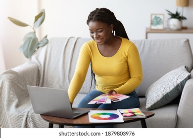 Beautiful Black Woman Interior Designer Holding Color Swatch Samples And Working On Laptop At Home, Checking Palette For New Design Project, Sitting On Sofa In Living Room, Choosing Gamma For Overhaul