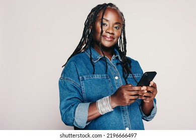 Beautiful Black Woman Looking At The Camera While Holding A Smartphone And Standing Against A Grey Background. Mature Woman With Dreadlocks Wearing A Denim Jacket And Make-up In A Studio.