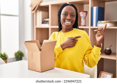 Beautiful Black Woman Opening Cardboard Box Smiling And Looking At The Camera Pointing With Two Hands And Fingers To The Side. 