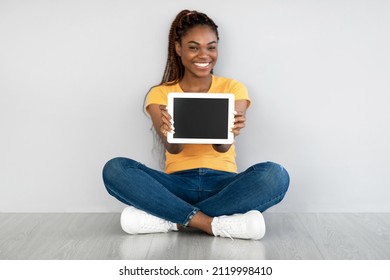 Beautiful Black Woman Showing Tablet Computer With Empty Screen, Sitting On Floor Against Grey Wall, Mockup For Website Or Application. African American Lady Demonstrating Touch Pad Display Template