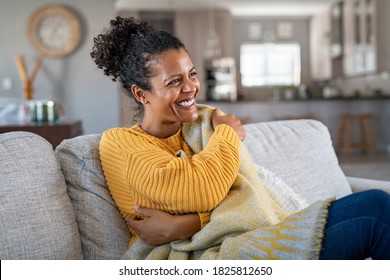 Beautiful black woman sitting on couch wrapped under blanket and laughing. Cheerful african american woman relaxing at home. Carefree and happy mid lady hugging herself with warm blanket in winter.