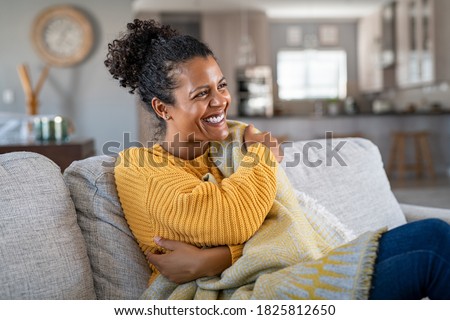 Beautiful black woman sitting on couch wrapped under blanket and laughing. Cheerful african american woman relaxing at home. Carefree and happy mid lady hugging herself with warm blanket in winter.
