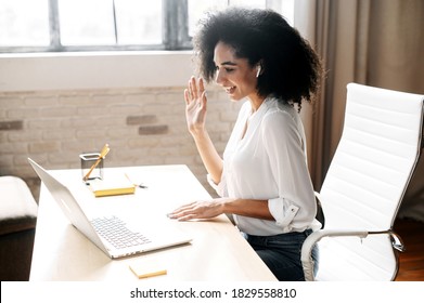 A beautiful black woman in smart casual wear is using laptop computer for video call at a modern office, a cheerful female employee is greeting colleagues and waving into webcam, side view - Shutterstock ID 1829558810