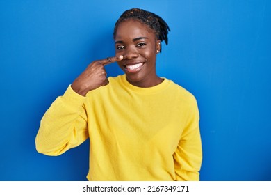 Beautiful Black Woman Standing Over Blue Background Pointing With Hand Finger To Face And Nose, Smiling Cheerful. Beauty Concept 