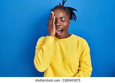 Beautiful Black Woman Standing Over Blue Background Yawning Tired Covering Half Face, Eye And Mouth With Hand. Face Hurts In Pain. 