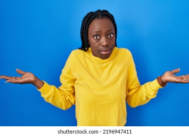 Beautiful Black Woman Standing Over Blue Background Clueless And Confused With Open Arms, No Idea Concept. 