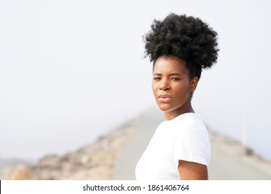 Beautiful Black Woman With Updo Afro Stands On The Jetty In The Fog At The Ocean Wearing A White Shirt And Looks Peacefully Towards The Camera                   