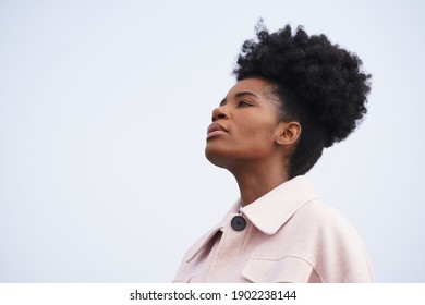 Beautiful Black Woman Wearing A Pink Jacket Stands Against A Stormy Blue Sky And Looks To The Left And Up In A Powerful Pose With And Updo Pony Tail Afro Hairstyle                              