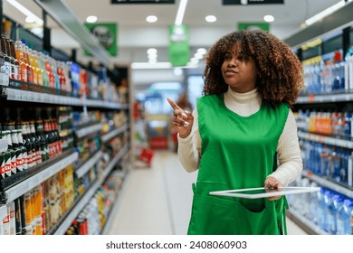 Beautiful black woman working in a supermarket. Female assistant using a digital tablet for taking inventory of products in display rack in a grocery store. - Powered by Shutterstock