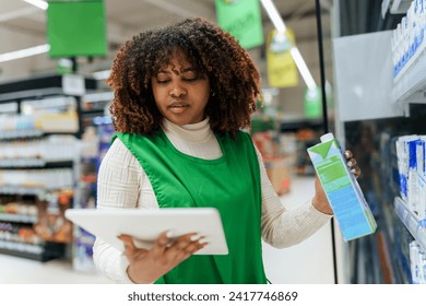 Beautiful black woman working in a supermarket. Female assistant using a digital tablet for taking inventory of products in display rack in a grocery store. - Powered by Shutterstock