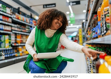 Beautiful black woman working in a supermarket. Female assistant using a digital tablet for taking inventory of products in display rack in a grocery store. - Powered by Shutterstock