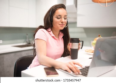 Beautiful Black-haired Woman Works From Home. An Employee Sits In The Kitchen And Has A Lot Of Work On A Laptop And Tablet And Has Video Conferencing And Meetings.