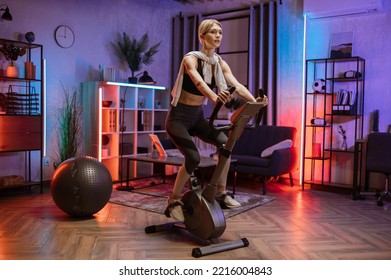 Beautiful blond caucasian sports women in sportswear cycling bike at home at night time. Cardio training, exercising legs, cardio workout indoors. - Powered by Shutterstock