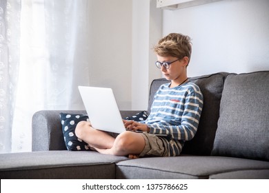 Beautiful Blond Child Sitting Legs Crossing Couch Home Using Computer. Young Boy Relaxing With Laptop Free Time Online Games. Teen Working Chat With Internet Friends. Student Doing Homework On The Web