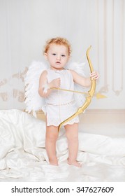 Beautiful blond curly toddler in a while lace dress with wings as a Cupid staying on the silk in the white room with gold moldings with golden bow in his hands. Saint Valentines Day celebration.  
