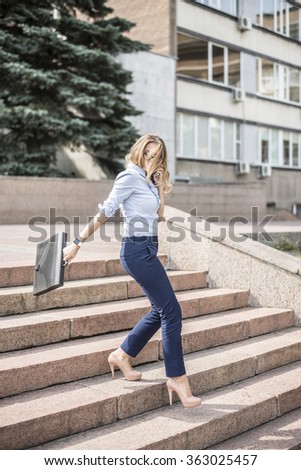 Beautiful blond female business woman walks in city park on stairs- carrying brief case over arm and talking on cell phone against summer trees