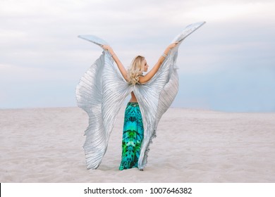 A beautiful blond girl in a golden with green dress with wings, suit is dancing an oriental, East dance in the desert on the sand. Beautiful exotic woman dancer in a costume for belly dancing.