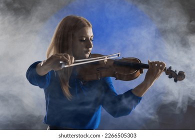 Beautiful blond girl playing violin. Female violinist against dark blue background with smoke.