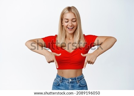 Beautiful blond girl pointing fingers down and looking at announcement with cute smile, showing advertisement, white background