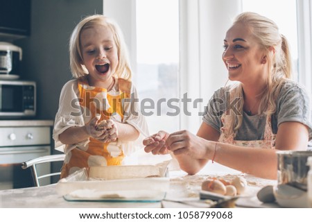 Beautiful blond mom teaching her daughter cooking on the kitchen. Parent making everyday breakfast together with child. Family at home lifestyle photo.