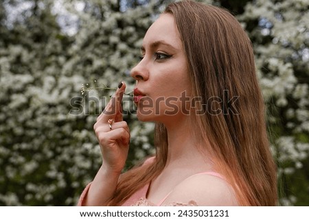 beautiful blond natural hair woman in pink outfit is posing in botanical garden park near blooming tree with flowers. Lady hold flower near mouth trying to taste and smell scent. Pretend to be smoking